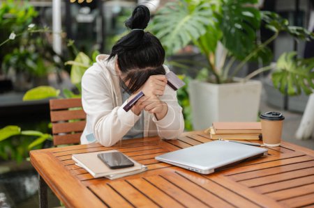 A stressed, upset Asian woman sitting at an outdoor cafe table, hand covering her face while holding a credit card, having financial problems or bankrupt.