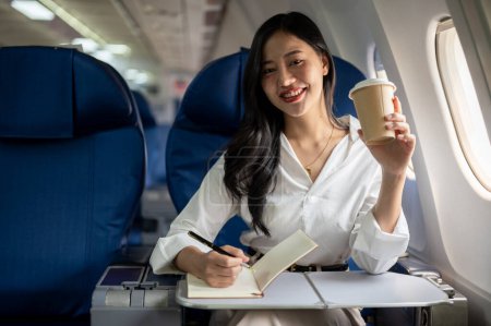 Téléchargez les photos : Une belle femme d'affaires asiatique sourit à la caméra alors qu'elle est assise à un siège de fenêtre dans un avion, voyageant pour un voyage d'affaires. Entrepreneurs et concepts de transport - en image libre de droit