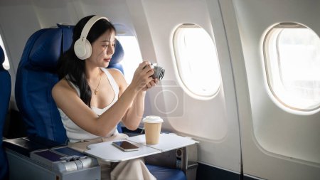 Téléchargez les photos : Une belle jeune femme asiatique est assise à une fenêtre dans un avion, prenant des photos avec son appareil photo pendant le vol, voyageant pour ses vacances d'été. style de vie et concepts de voyage - en image libre de droit