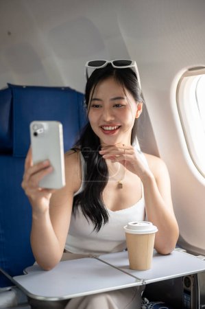Téléchargez les photos : Une femme asiatique séduisante et joyeuse prend un selfie ou enregistre une vidéo d'elle-même avec son smartphone pendant le vol pour son voyage de vacances, voyageant en avion. - en image libre de droit