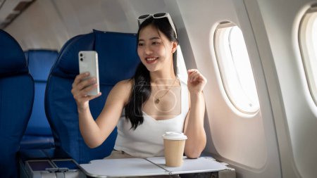 Téléchargez les photos : Une femme asiatique séduisante et joyeuse prend un selfie ou enregistre une vidéo d'elle-même avec son smartphone pendant le vol pour son voyage de vacances, voyageant en avion. - en image libre de droit