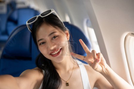 Téléchargez les photos : Une jolie voyageuse asiatique montre un signe de paix avec deux doigts tout en prenant un selfie ou en enregistrant un vlog de voyage d'elle-même pendant le vol pour son voyage de vacances d'été. - en image libre de droit