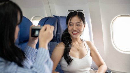 Téléchargez les photos : Une belle jeune femme asiatique est photographiée par son amie pendant le vol, voyageant ensemble en avion pour le voyage d'été. classe économique, tourisme, voyage de filles, transport - en image libre de droit