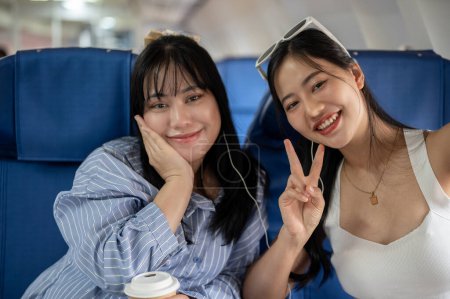 Téléchargez les photos : Deux femmes asiatiques heureuses montrent un signe de paix avec deux doigts tout en prenant un selfie ensemble pendant le vol pour leur voyage de vacances d'été. voyage de filles, touriste, voyageur, amitié - en image libre de droit
