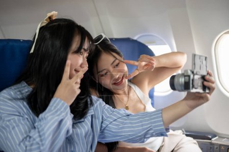 Téléchargez les photos : Deux femmes asiatiques heureuses montrent un signe de paix avec deux doigts tout en prenant un selfie ensemble pendant le vol pour leur voyage de vacances d'été. voyage de filles, touriste, voyageur, amitié - en image libre de droit