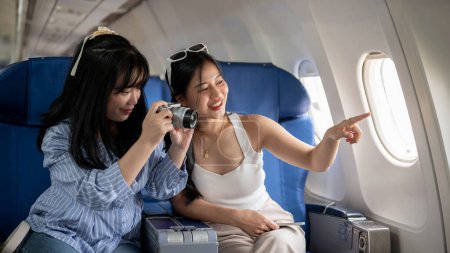 Téléchargez les photos : Deux jeunes amies asiatiques joyeuses aiment parler et prendre des photos pendant le vol, voyageant en vacances ensemble en avion. amitié, mode de vie, concepts de transport - en image libre de droit
