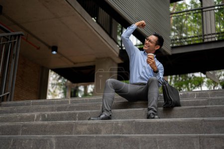 A happy, joyful Asian businessman sits on steps outside a building, holding a coffee cup and cheering with a raised fist, showing joy and successful expression.