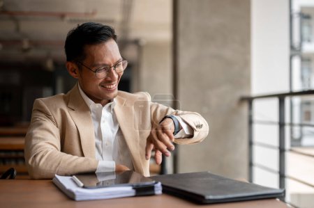 Ein selbstbewusster, lächelnder asiatischer Millennial-Geschäftsmann in beigem Anzug und Brille kontrolliert die Zeit an seiner Armbanduhr, während er an einem Tisch in einem Hausflur sitzt..