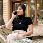An attractive Asian woman sits at an outdoor table in a garden of a cafe talking on the phone with someone. people, lifestyle, and wireless technology concepts