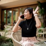 A beautiful, positive Asian woman sits at an outdoor table of a cafe talking on the phone while waving her hand to call or greet someone. people and lifestyle concepts