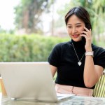 An attractive Asian woman or businesswoman in casual clothes sits at a table in a green garden or backyard using her laptop computer, talking on the phone, working remotely.