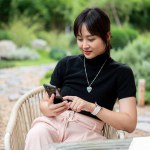 A beautiful Asian woman in casual clothes sits at a table in a beautiful green backyard relaxing and using her phone. people, lifestyle, and wireless technology concepts