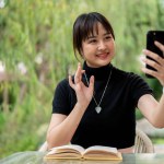 A cheerful Asian woman in casual clothes is talking on a video call on her smartphone with someone while relaxing at a table in a green garden backyard. people and wireless technology concepts