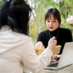 An attractive Asian woman in casual clothes working remotely in a garden with her colleague, discussing work and sharing ideas.