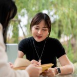 An attractive Asian woman in casual clothes working remotely in a garden with her colleague, discussing work, sharing ideas, and explaining some details in the book.