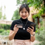 A beautiful, positive Asian woman holding a credit card and a smartphone while walking in a beautiful garden. mobile banking, credit card or debit card payment, cashless