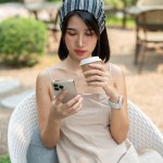 A beautiful Asian woman in a cute minimal dress sits in a beautiful English garden sipping coffee and using her smartphone. chatting, social media, mobile app