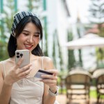 An attractive, smiling Asian woman in a cute minimal dress sits at a table in a beautiful English garden holding a credit card and smartphone, enjoying shopping online. mobile banking, online payment
