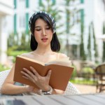 A beautiful Asian woman in a cute dress relaxes in a beautiful garden on a bright day, reading a book. leisure, hobby, lifestyle