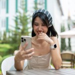 An attractive Asian woman in a cute dress laughs at her phone and enjoys chatting with her friends online while relaxing in a garden on a bright day.