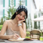 An attractive Asian woman in a cute dress writing her diary at a table in a beautiful garden on a bright day. studying, keeping diary, making list, working
