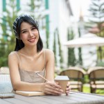 A beautiful, attractive Asian woman sits at a table with a book and a coffee cup on the table in a beautiful garden on a bright day, smiling at the camera.