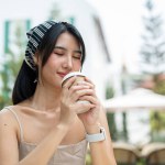 An attractive Asian woman in a cute dress smells her coffee, eyes closed, enjoying her coffee while relaxing in a beautiful garden on a bright day.