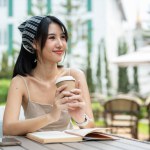 An attractive Asian woman in a cute dress is holding a coffee cup, looking away from the camera, and thinking or daydreaming about something while sitting at a table in a beautiful garden.