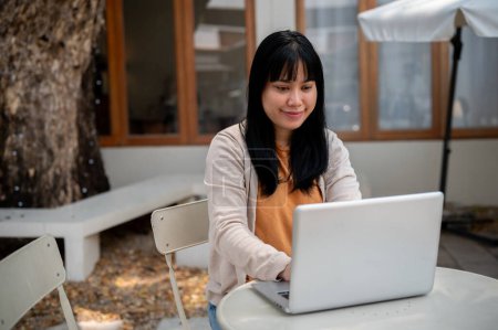A positive young Asian woman in comfy clothes is working remotely at an outdoor table of a cafe, focusing on her project or responding to emails on her laptop computer.