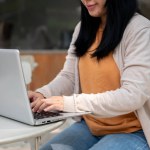 A cropped image of a woman in comfy clothes responds to emails, types on her laptop keyboard, and works remotely on her laptop at an outdoor table of a cafe on a bright day.