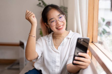 A young, cheerful Asian woman shows her clenched fist in a triumphant pose, celebrating good news on her smartphone indoors. people and wireless technology concepts