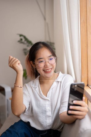 A young, cheerful Asian woman shows her clenched fist in a triumphant pose, celebrating good news on her smartphone indoors. people and wireless technology concepts