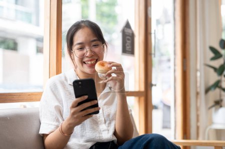 A young, happy Asian woman enjoying a donut while using her smartphone on a sofa in a cafe. reading online blogs, chatting, watching videos on social media, shopping online, using a mobile app