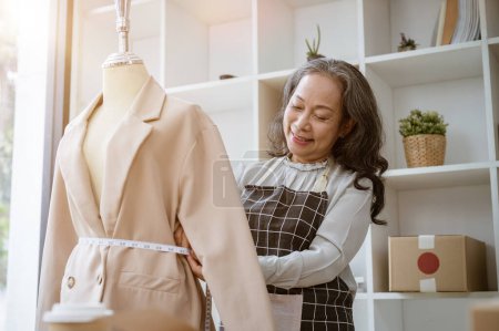 An attractive, experienced mature Asian female suit maker or tailor is measuring and adjusting the suit pattern on a dummy, working in her atelier studio. fashion designer, garment business