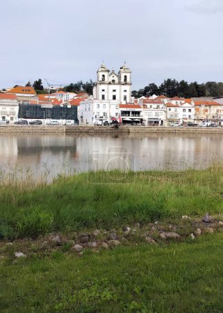 Blick auf das historische Zentrum der Stadt über den Fluss Sado, im Vordergrund grasbewachsenes linkes Ufer des Flusses, Alcacer do Sal, Portugal - 6. März 2024