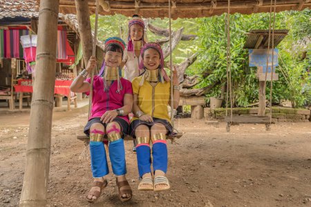 Foto de Karen mujeres de Pai en Mae Hong Son, Tailandia, con anillos de cuello de latón, se sienta en el columpio de madera en la aldea rústica, vistiendo una vibrante blusa de color, cubiertas vibrantes de patas de color y un colorido abrigo para la cabeza - Imagen libre de derechos