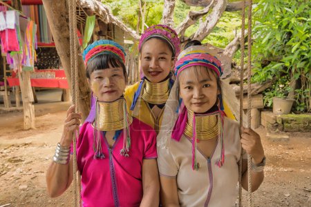 Foto de Karen mujeres de Pai en Mae Hong Son, Tailandia, con anillos de cuello de latón, se sienta en el columpio de madera en la aldea rústica, vistiendo una vibrante blusa de color, cubiertas vibrantes de patas de color y un colorido abrigo para la cabeza - Imagen libre de derechos