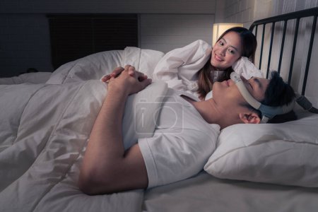 Foto de Feliz pareja asiática con la máquina CPAP en la mesa auxiliar con el marido con máscara CPAP durmiendo suavemente en la cama toda la noche sin roncar - Imagen libre de derechos