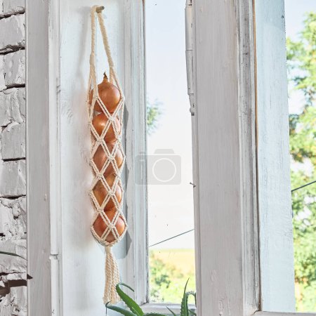 Onions hang on the wall in nets. Vegetables keeper macrame. Kitchen neutral apartment wall decor.