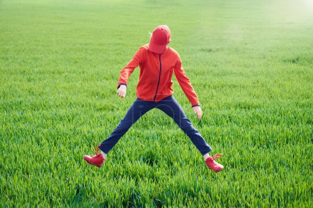 A teenager is jumping with his legs apart against the background of a green grass field.