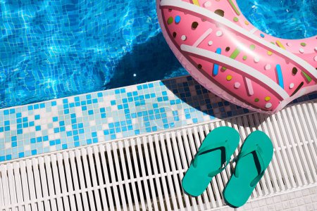 Anillo inflable rosa y chanclas de goma verde junto al agua azul de la piscina al aire libre. Relajación junto a la piscina.