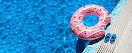 Pink inflatable ring and rubber flip-flops by blue outdoor pool water. Poolside relaxation.