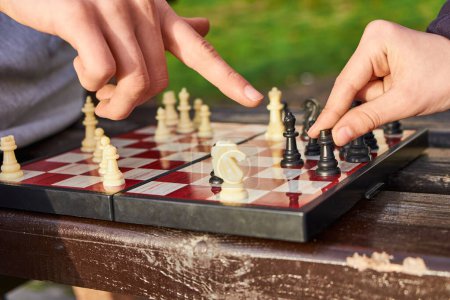 Person, die Schach in der Natur spielt. Die Hand eines Schachspielers schwebt über einem Turm, gerade dabei, einen Zug zu machen. Die Hand eines anderen Spielers zeigt auf das Brett und bietet einen Vorschlag.