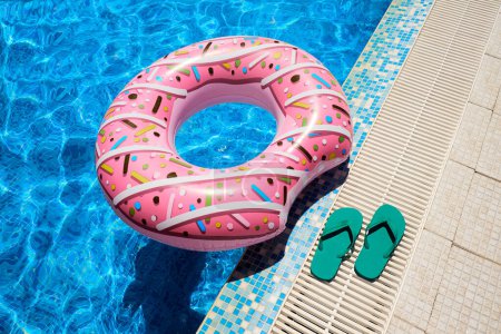 Pink inflatable ring and green rubber flip-flops by blue outdoor pool water. Poolside relaxation.