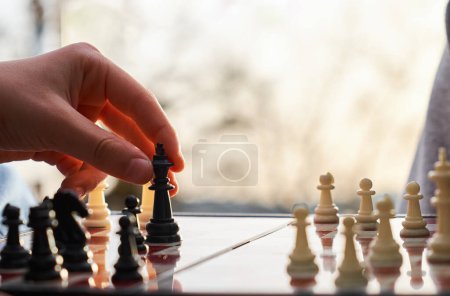 The chess board is made of wood. Person playing chess in nature. The person is holding a black king in their hand.