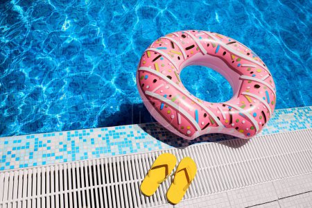 Pink inflatable ring and yellow rubber flip-flops by blue outdoor pool water. Poolside relaxation.