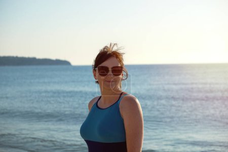 Una mujer madura y segura disfruta de un día en la playa, con un traje de baño cerrado y gafas de sol.
