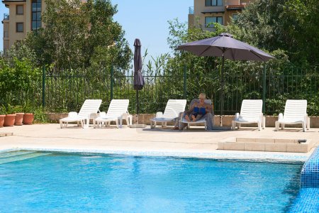Livre de lecture adolescent solitaire, détente sur une chaise longue au bord de la piscine bleue près de l'hôtel.