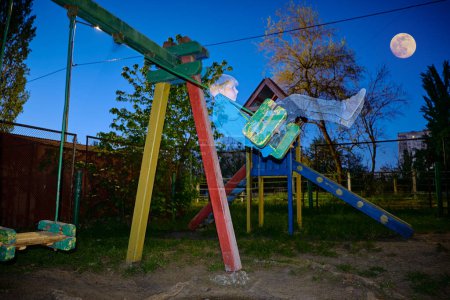 Abandoned playground with a transparent boy swinging on a swing set in the moonlight.