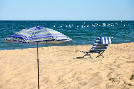 Scène de plage relaxante avec une chaise de plage rayée bleue et blanche et un parapluie rayé bleu et vert sous un ciel bleu clair avec des nuages blancs et un troupeau de mouettes volant au-dessus.